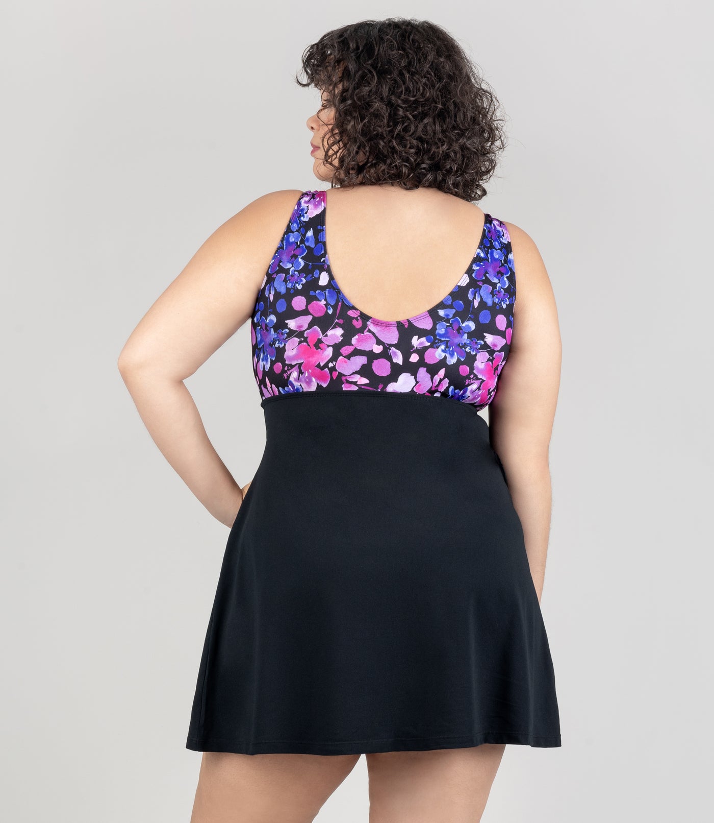 JunoActive model wearing Aquasport Shirred Swim Dress. Top of dress is pink and blue floral elegance print and under bust is black fabric. Model facing back.