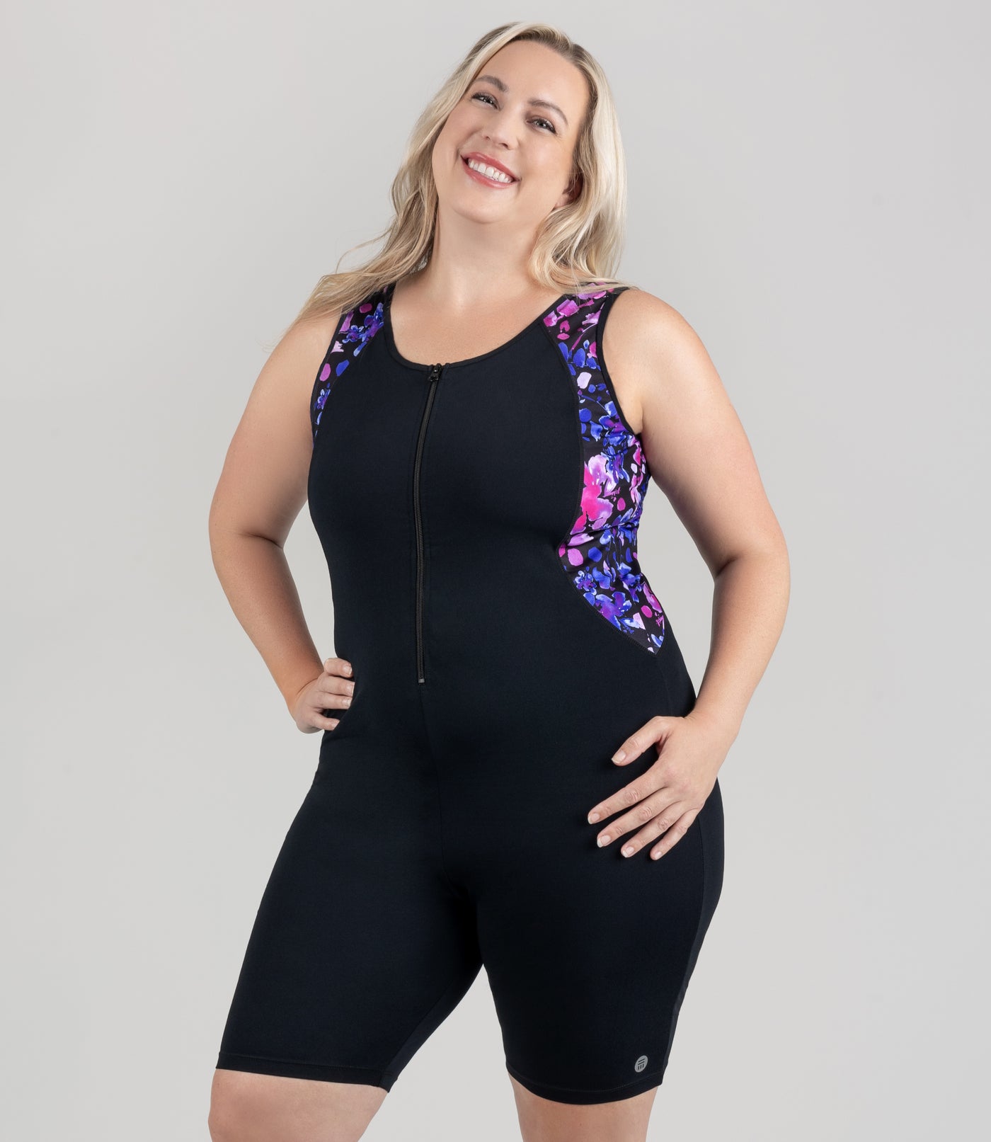 Model is wearing JunoActive's Aquasport Crossback Aquatard. She's facing front with her  hands on her hips. The suit has a strip on the side of pink and blue floral elegance print from the shoulders and end just to the top of the hip bone.
