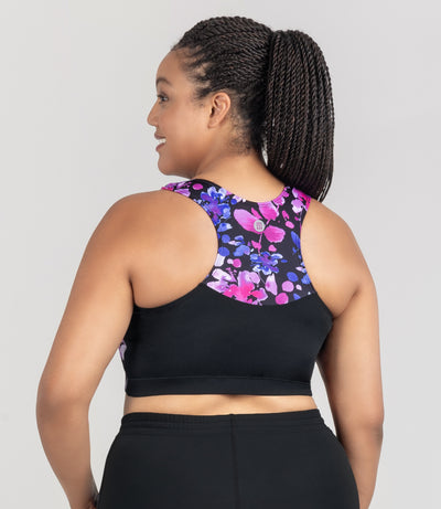 JunoActive model, facing back, wearing Aquasport Hanalei Bra Top featuring blue and pink floral elegance print at top back of bra and solid black on bottom half back of bra. Model facing front with her left hand resting on her left leg. Right arm is to her right side. 