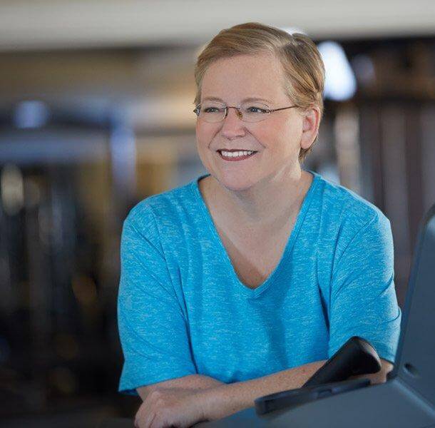 Anne Kelly, JunoActive founder and president, leaning against exercise equipment. She is looking off to her right, smiling and is wearing a blue JunoActive plus size short sleeve activewear top.  The background is a blurred gym.