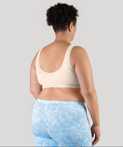 Plus size woman, facing back, wearing JunoActive Stretch Naturals Shirred Bra Top. Bra is gathered in middle and is a scoop top neck. Straps are wide and bra color is sandy.