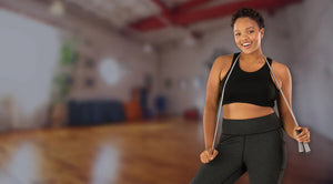 African American plus size woman with black JunoActive plus size sports bra in the gym. She is smiling at the camera and has a jumprope wrapped around her neck.