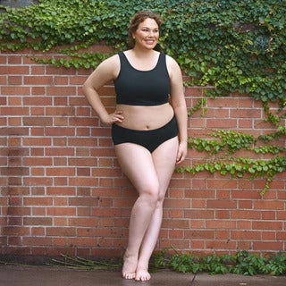 Plus size woman leaning against a brick wall with her hand on her hip.  She is wearing a black JunoActive plus size bra and a black JunoActive plus size brief. The background is a brick wall with green vines growing on the wall.