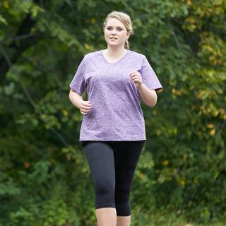 Plus size woman facing front while jogging. She is wearing a purple JunoActive short sleeve plus size activewear top and black JunoActive plus size capri length leggings. The background is green trees