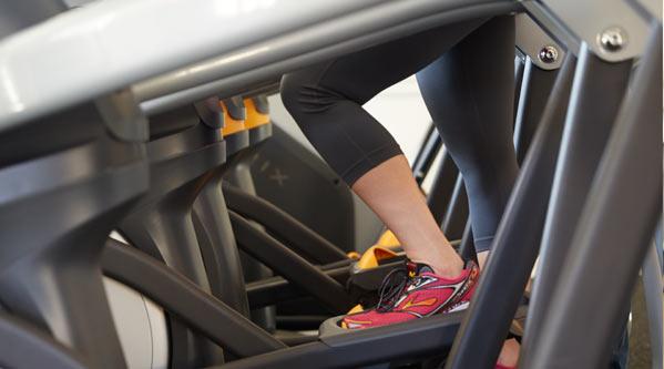 The bottom of multiple stair climber exercise machines with a woman's legs and feet exercising on one of the machines. She is wearing black JunoActive QuikWik plus size capri leggings and red sneakers.