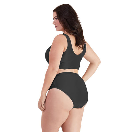 Plus size woman, facing back left, wearing JunoActive plus size QuikWik Soft Control Bra Top in Black. The woman is wearing a pair of coordinating QuikWik Comfort plus size Briefs in Black. 