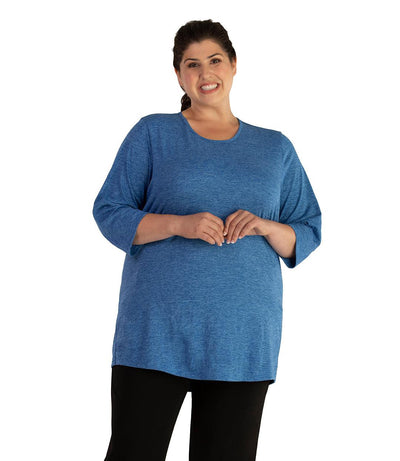 Plus size woman, facing front, wearing JunoActive plus size QuikLite Scoop Neck ¾ Sleeve Top in the color Heather Blue.  She is wearing JunoActive Plus Size Leggings in the color Black.