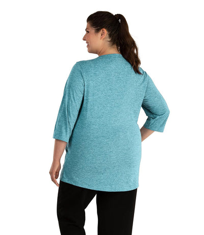Plus size woman, facing back looking left, wearing JunoActive plus size QuikLite Scoop Neck ¾ Sleeve Top in the color Heather Deep Teal. She is wearing JunoActive Plus Size Leggings in the color Black.