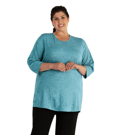 Plus size woman, facing front, wearing JunoActive plus size QuikLite Scoop Neck ¾ Sleeve Top in the color Heather Deep Teal. She is wearing JunoActive Plus Size Leggings in the color Black.