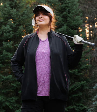 Plus size woman wearing JunoActive's JunoStretch Mock Neck Jacket in color black with warm mauve lining cuffs and warm mauve piping details along shoulders and pockets. She is wearing a golf glove and visor and is holding a golf club over one shoulder and looking off to her right. Tall evergreen trees are in the background.
