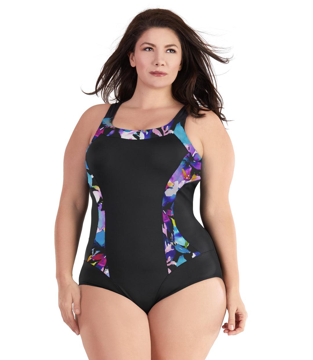 Plus size woman, facing front, wearing JunoActive plus size QuikEnergy Color Block Tank Suit South Pacific Black. The blocking at the neckline and princess panels is a multi colored watercolor floral print. Side panels, straps, center front, and leg blocking are black, has a scoop neckline and conservative leg opening.