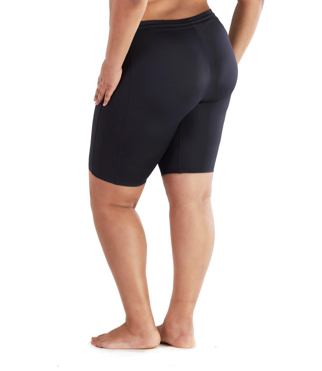 Bottom half of plus size woman facing back wearing JunoActive QuikEnergy Fitted Swim Short. The short is black and is tight to the body. The hem is just above the knee.