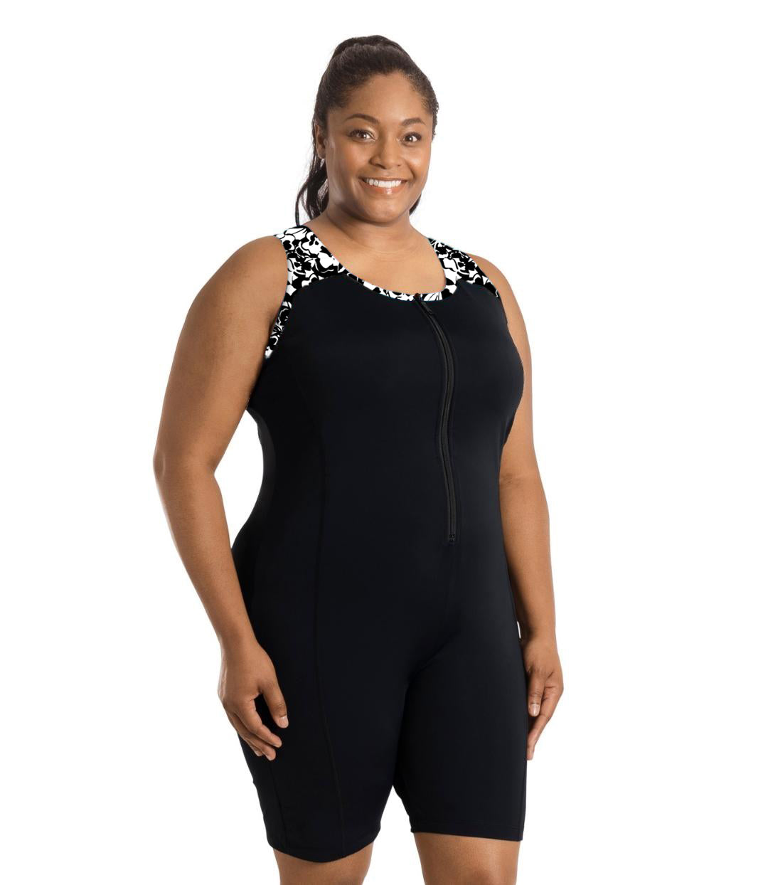 Plus size woman, facing front, wearing JunoActive plus size QuikEnergy Racerback Zip Front Aquatard Hibiscus print. Neck binding, armhole binding, and front shoulder blocking is a black and white print. The main body of the suit is solid black, has a black zipper in front. The leg hits a few inches above the knee.