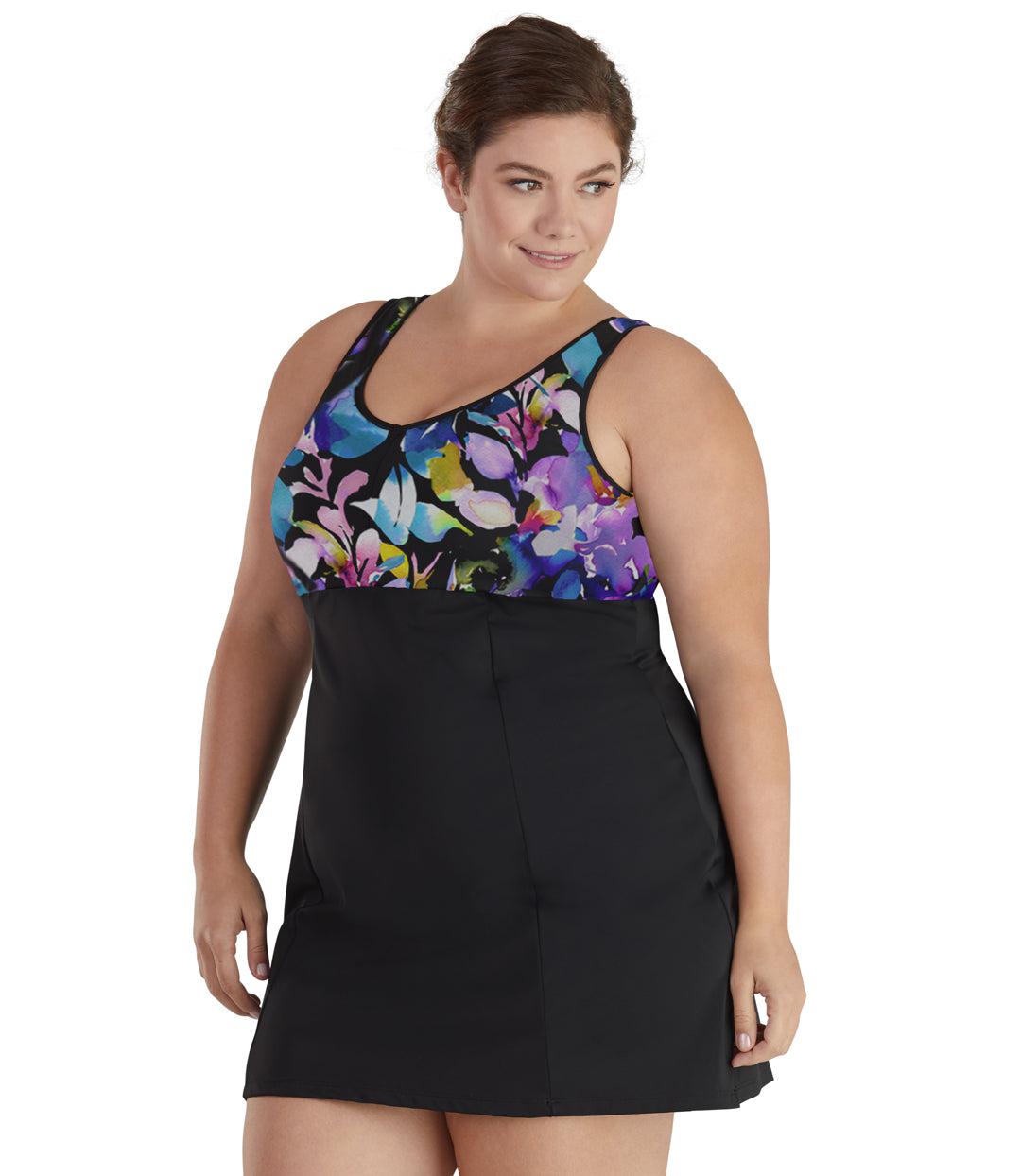 Plus size woman, facing front, wearing JunoActive plus size QuikEnergy Swim Dress South Pacific Black. The top of swim dress is a multi colored watercolor floral print. The bottom of the dress is solid black, has a slight a-line shape and ends mid-thigh.