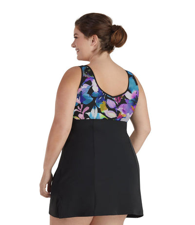 Plus size woman, back view, wearing JunoActive plus size QuikEnergy Swim Dress South Pacific Black. The top of swim dress is a multi colored watercolor floral print. The bottom of the dress is solid black, has a slight a-line shape and ends mid-thigh.