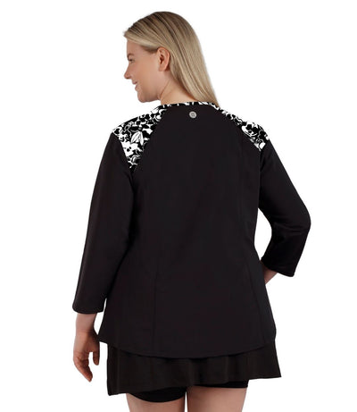 Plus size woman, facing back, wearing QuikEnergy ¾ Sleeve Swim and Sun Top Black and hibiscus print on shoulders and solid black colorblocking on the sleeves and torso.