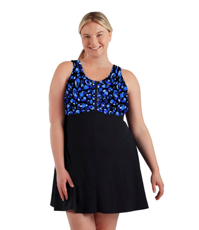 Plus size woman, facing front, wearing JunoActive plus size AquaSport Zip Front Swim Dress Ocean Blues Print Black. The top of the swim dress has a multi colored blue bubble print and front zipper. The bottoms of the dress is solid black, has a slight a-line shape and ends mid-thigh.