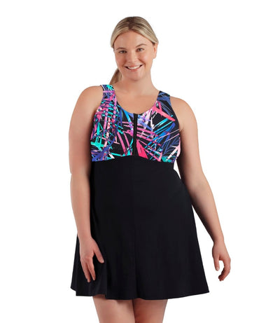 Plus size woman, facing front, wearing JunoActive plus size AquaSport Zip Front Swim Dress Sunset Palm Print Black. The top of the swim dress has a multi colored pink blue and black Palm print and front zipper. The bottoms of the dress is solid black, has a slight a-line shape and ends mid-thigh.