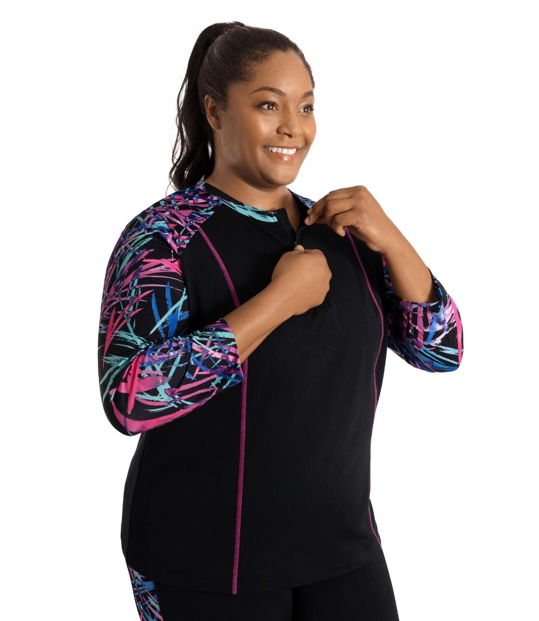 Plus size woman, side facing view, wearing AquaSport Long Sleeve Rash Guard Sunset Palms Print Black. Quarter zip closure detail at neck. Colorblock pink, blue and teal abstract line print on three quarter length sleeves and pink princess seams.