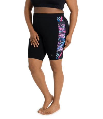 Bottom half of plus sized woman, facing front, wearing JunoActive AquaSport Long Fitted Swim Shorts Sunset Palms Print Black. Pink, blue, teal abstract line print colorblock accent on the side of the leg. JunoActive Detail near the hem of the short. 