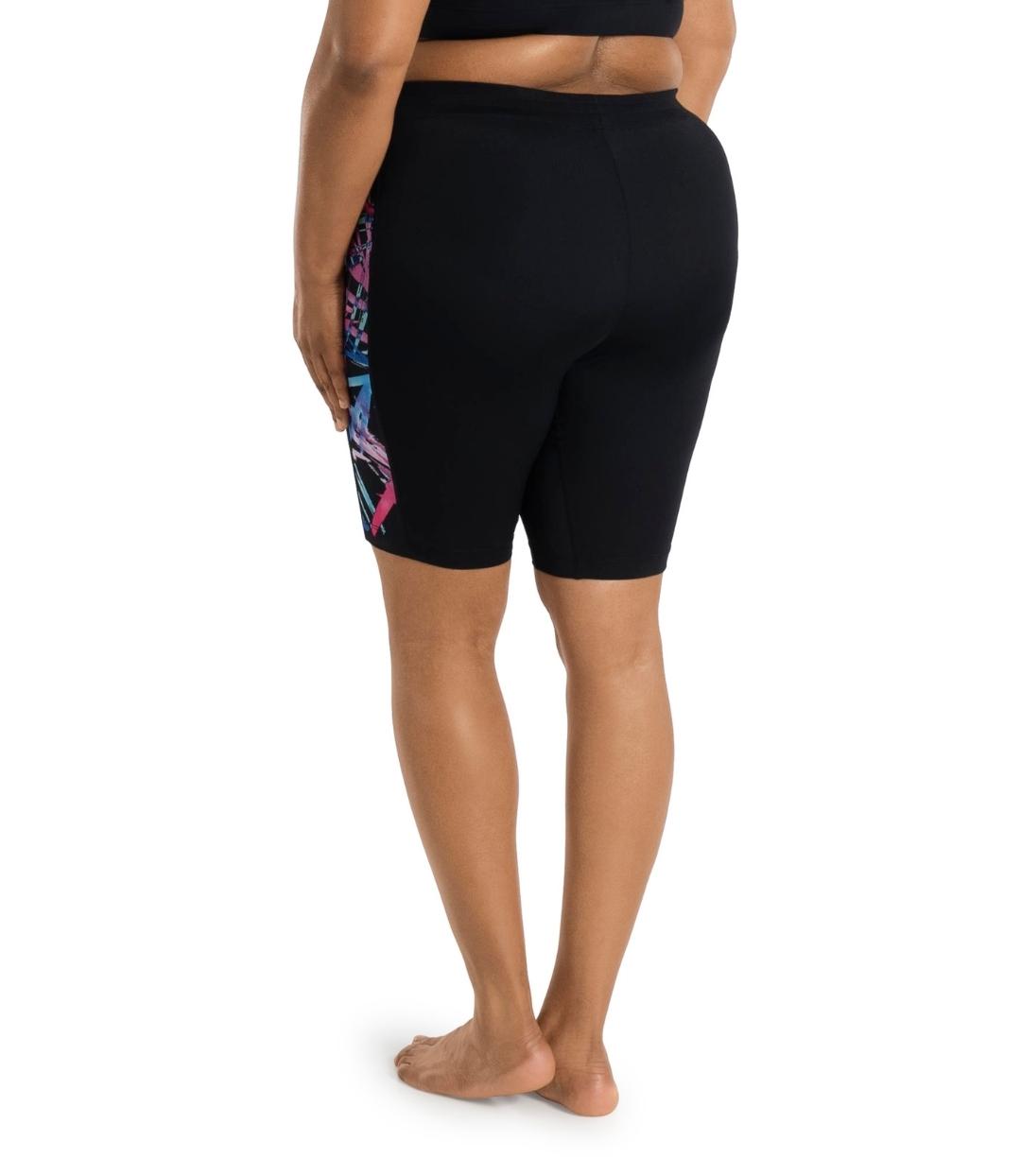 Bottom half of plus sized woman, back view, wearing JunoActive AquaSport Long Fitted Swim Shorts Sunset Palms Print Black.The shorts have pink, blue and teal abstract line print colorblock accent on the side of the leg. 