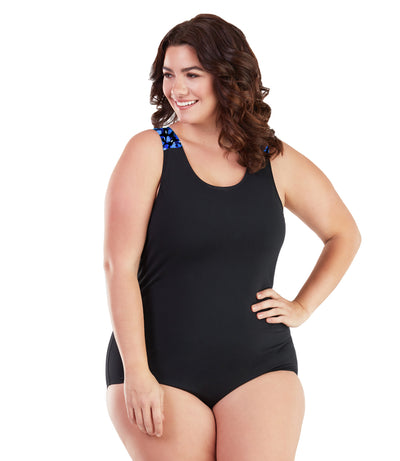 Plus size woman, facing front, wearing JunoActive plus size AquaSport Crossback Tanksuit Ocean Blues Print Black. The shoulders of the tanksuit has a multi colored blue bubble print blocking. The main body of the suit is solid black, has a scoop neckline and conservative leg opening.