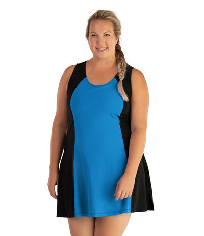 Plus size woman, facing front, wearing JunoActive plus size AquaSport Crossback Swim Dress Pacific Blue Black. The center front of the dress is blue. The sides of the dress is solid black, has a slight a-line shape and ends mid-thigh.