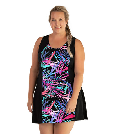 Plus size woman, facing front, wearing JunoActive plus size AquaSport Crossback Swim Dress Sunset Palm Print Black. The center front of the dress has a multi colored pink blue and black Palm print. The sides of the dress is solid black, has a slight a-line shape and ends mid-thigh.