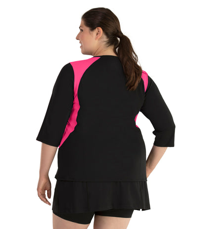 Plus size woman, facing back to side profile, wearing AquaSport Three Quarter Sleeve Rash Guard Pink and Black. V-neckline, black torso and sleeves with pink colorblock at shoulder and waist. She is wearing a pair of black JunoActive plus size swim shorts.