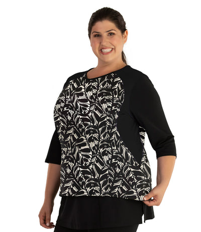 Plus size woman, facing front, wearing AquaSport Three Quarter Sleeve Rash Guard Tropical Print. V-neckline,   black tropical print torso and black colorblocked sleeves and side of waist.   She is wearing a pair of black JunoActive plus size swim shorts. 