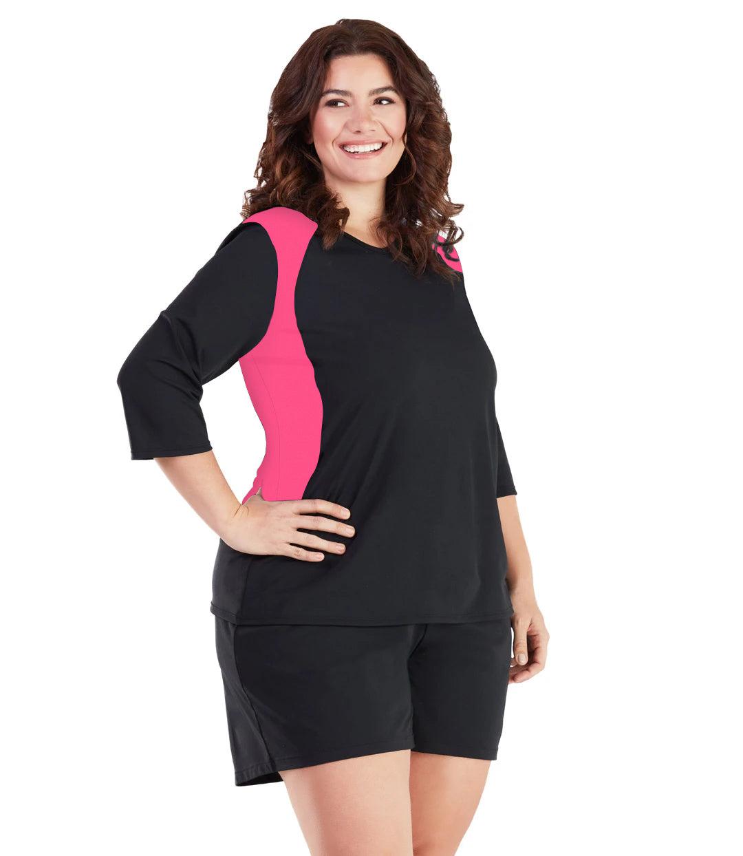 Plus size woman, facing front, wearing AquaSport Three Quarter Sleeve Rash Guard Pink and Black. V-neckline, black torso and sleeves with pink colorblock at shoulder and waist. She is wearing a pair of black JunoActive plus size swim shorts. 