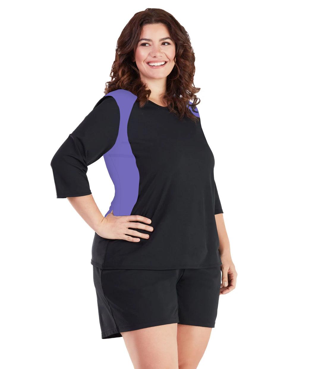 Plus size woman, facing front, wearing AquaSport Three Quarter Sleeve Rash Guard Purple and Black. V-neckline,  black torso and sleeves with purple colorblock at shoulder and waist.   She is wearing a pair of black JunoActive plus size swim shorts. 
