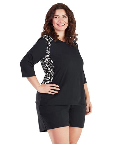 Plus size woman, facing front, wearing AquaSport Three Quarter Sleeve Rash Guard Tropical Black. V-neckline,  black torso and sleeves with tropical print colorblock at shoulder and waist.   She is wearing a pair of black JunoActive plus size swim shorts. 