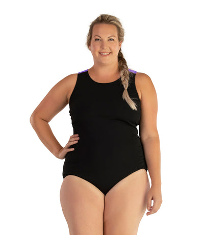 Plus size woman, facing front, wearing JunoActive plus size AquaSport Crossback Tanksuit Purple Black. The shoulders have purple color blocking. The main body of the suit is solid black, has a scoop neckline and conservative leg opening.