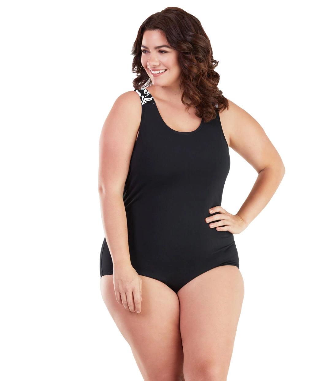 Plus size woman, facing front, wearing JunoActive plus size AquaSport Crossback Tanksuit Tropical Print Black. The shoulders of the tanksuit has a Black and white Tropical leaf print. The main body of the suit is solid black, has a scoop neckline and conservative leg opening.