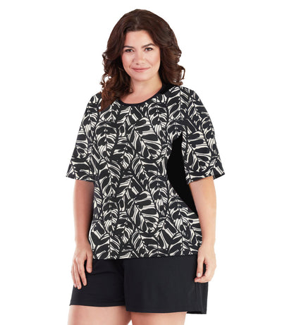 Plus size woman, facing front, wearing AquaSport Colorblock Swim Tee Tropical Print. Black colorblocking on waist and shoulders. Long cover short sleeves meeting at the elbow and a rounded neckline. 