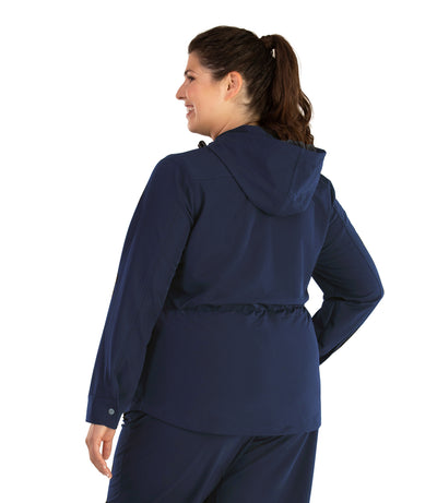 Plus size woman, back view , wearing a JunoActive plus size Hiking and Travel Jacket in navy blue. The jacket has a cinched waist, hood, zip front closure, and side zip pockets. The length of the plus size jacket hits at the hips.