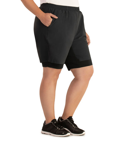 Bottom half of plus sized woman, facing to side, hand in pocket, wearing JunoActive Dual Layer Walking Short in black. These shorts have two layers with the bottom being tight bike short and the outer is shell like material and looser fitted. The hem is above knees.