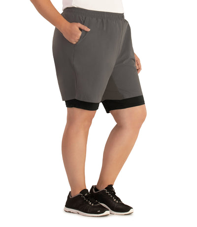 Bottom half of plus sized woman, facing to side, hand in pocket, wearing JunoActive Dual Layer Walking Short in black and oak gray. These shorts have two layers with the bottom being a black tight bike short and the outer is shell is like material and looser fitted in oak gray. The hem is above knees.