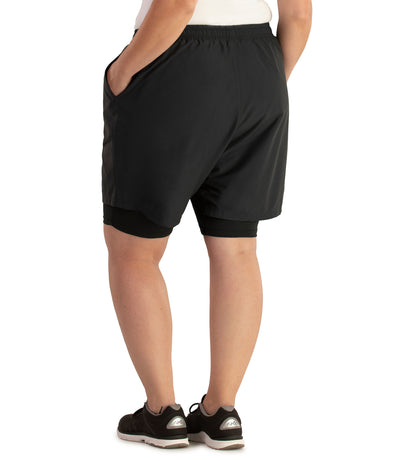 Bottom half of plus sized woman, facing with back to viewer, hands in pockets, wearing JunoActive Dual Layer Walking Short in black. These shorts have two layers with the bottom being tight bike short and the outer is shell like material and looser fitted. The hem is above knees.
