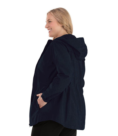 Plus size woman, side view, facing left wearing a JunoActive plus size Waterproof Breathable Wind & Rain Jacket in deep navy. The jacket has a hood, double front closure and side pockets. The length of the plus size jacket hits just below the hips.