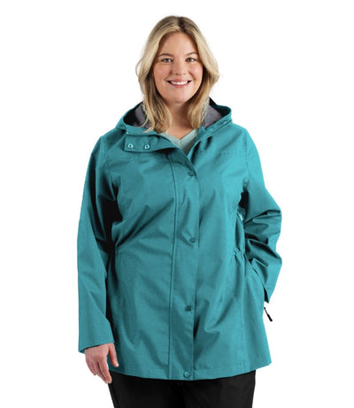 Plus size woman, front view, wearing a JunoActive plus size wind and rain jacket in malechite blue. The jacket has a hood, double front closure and side pockets. The length of the plus size jacket hits just below the hips.