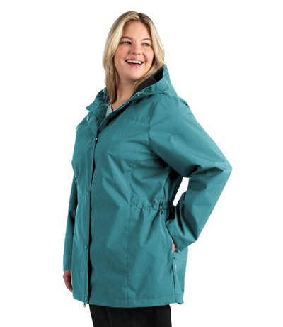 Plus size woman, side view, looking over her shoulder wearing a JunoActive plus size wind and rain jacket in malechite blue. The jacket has a hood, double front closure and side pockets. The length of the plus size jacket hits just below the hips.