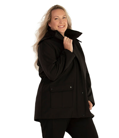 Plus size woman, side view facing right, wearing a JunoActive plus size Hooded Softshell Jacket in Black. The jacket has a removeable hood, zip front closure, zip chest pocket, and snap patch pockets at the hip. The length of the plus size jacket hits below the hips.