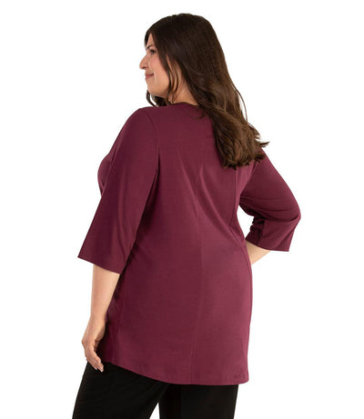 Plus size woman, facing back looking left, wearing JunoActive plus size Stretch Naturals Lite ¾ Sleeve Swing Top in the color Dark Plum. She is wearing JunoActive Plus Size Leggings in the color Black.