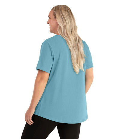 Plus size woman, facing back left, wearing JunoActive plus size Stretch Naturals Lite Short Sleeve Scoop Neck Top in the color Mountain Lake Blue. She is wearing JunoActive Plus Size Leggings in the color black. 