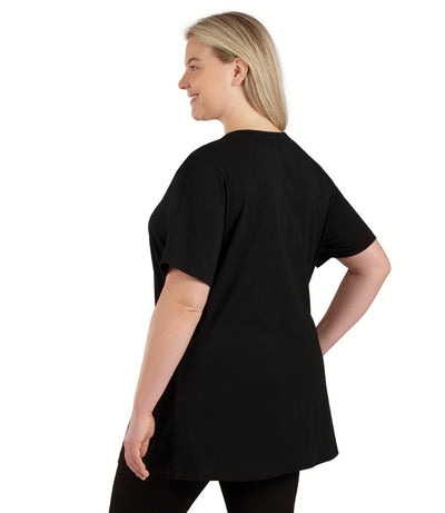 Plus size woman, facing back left, wearing JunoActive plus size Stretch Naturals Lite Short Sleeve Split Collar Tunic in the color Black. She is wearing JunoActive Plus Size Leggings in the color black. 