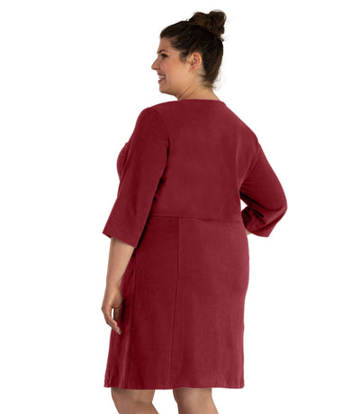 Plus size woman, facing back looking left, wearing JunoActive plus size Legacy Cotton Casual ¾ Sleeve Dress in the color Garnet. Her right hand is in the dress pocket at her hip and her left hand falls naturally at her side. The dress length is at her knee.