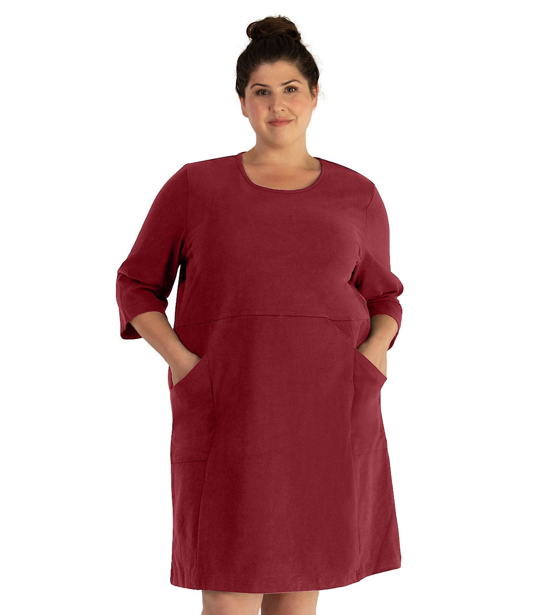 Plus size woman, facing front, wearing JunoActive plus size Legacy Cotton Casual ¾ Sleeve Dress in the color Garnet. Both hands are in the dress pockets at her hip. The dress length is at her knee. 