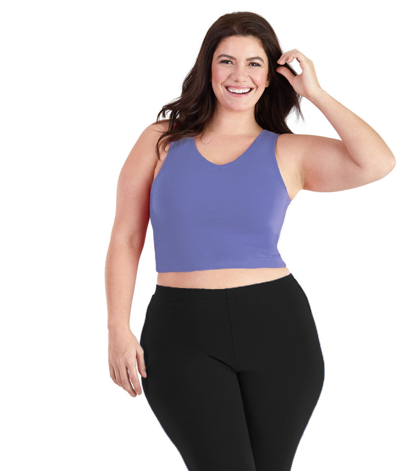 Plus size woman, facing front, wearing JunoActive plus size Stretch Naturals V-Neck Bra top in Cornflower Blue. The woman is wearing black JunoActive plus size leggings. Her left arm is tucking her hair behind her ear, her right arm falls naturally to her side. 
